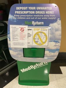 A green MedReturn drug collection unit with instructions and a warning sign. Text encourages depositing unwanted prescription drugs to protect children and water supply.