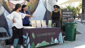 Three women stand near a UCF Arboretum information table with pamphlets. They are located next to a mural and a green waste container. One woman is pointing at the table while talking to the others.