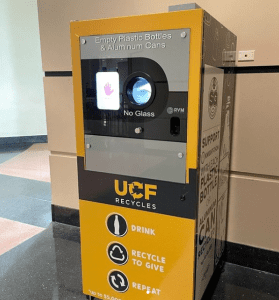 A UCF Recycles machine for collecting empty plastic bottles and aluminum cans, with a slot for depositing items and a screen displaying instructions. The station promotes recycling and sustainability.
