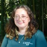 Person with glasses and curly hair wearing a blue shirt, standing outdoors with greenery in the background.