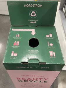 A Nordstrom BeautyCycle recycling collection box for beauty product waste. The box is green and pink with instructions for recyclable items like pumps, droppers, caps, and squeeze tubes.