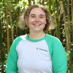 A person with short, wavy hair and a green and white "Arboretum" shirt smiles while standing in front of tall bamboo plants.