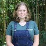 A person with shoulder-length hair and wearing a green shirt and blue overalls stands in front of a dense bamboo grove.