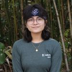 Person with glasses wearing a green long-sleeve shirt and black bandana standing in front of dense bamboo foliage.