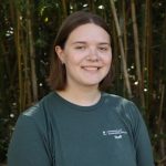 A person with short brown hair and a green staff shirt stands smiling in front of a bamboo forest.