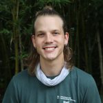 A person is smiling in front of a dense bamboo forest. They are wearing a green t-shirt and a neck scarf.