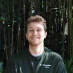 A person is smiling outdoors, wearing a green "Arboretum Intern" shirt. They're standing in front of a bamboo background.