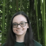 A person wearing glasses and a dark green shirt stands in front of green bamboo plants, smiling at the camera.