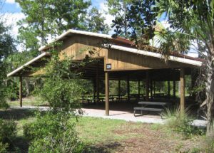 An open wooden pavilion with a picnic area enveloped by trees and shrubs, this tranquil spot offers a serene retreat. To fully enjoy the experience, ensure you secure a site use permit for your gathering.