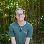 A person wearing glasses and a green staff t-shirt sits outdoors in front of tall bamboo and green plants, smiling at the camera.