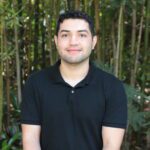 Carlos with short dark hair wearing a black polo shirt sits in front of tall green bamboo and foliage outdoors.