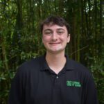 A person wearing a black polo shirt with a "Learning by Leading" logo stands outdoors in front of dense green bamboo foliage, smiling at the camera.
