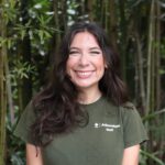 A woman wearing an "Arboretum Staff" t-shirt smiles while standing in front of tall green plants outdoors.