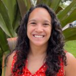 A woman with long curly hair wearing a red polka dot top smiles in front of large green leaves outdoors.
