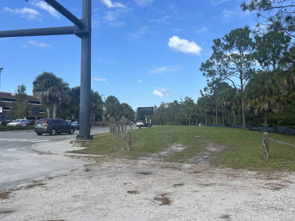 Grassy area beside a road with cars parked on the left, bordered by trees and wooden posts, under a mostly clear blue sky.