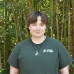 A person with short brown hair stands in front of tall green bamboo, wearing a dark green "Learning by Leading" t-shirt and a necklace with a stone pendant.