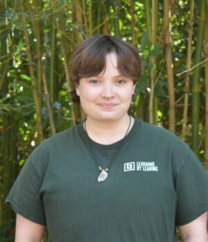 A person with short brown hair stands in front of tall green bamboo, wearing a dark green "Learning by Leading" t-shirt and a necklace with a stone pendant.