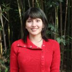 A person with short brown hair in a red sweater stands outdoors in front of tall green bamboo plants, smiling at the camera.