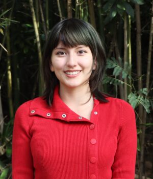 A person with short brown hair in a red sweater stands outdoors in front of tall green bamboo plants, smiling at the camera.