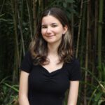 A young woman with long brown hair, wearing a black top, stands in front of dense green bamboo plants, smiling slightly at the camera.