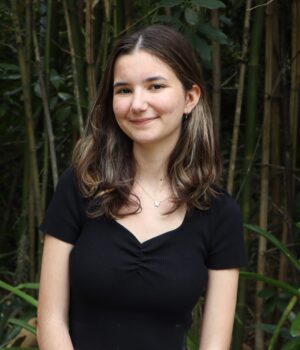 A young woman with long brown hair, wearing a black top, stands in front of dense green bamboo plants, smiling slightly at the camera.