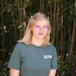 A person with blonde hair, glasses, and a green "Learning by Leaving" t-shirt stands outdoors in front of tall plants and bamboo.
