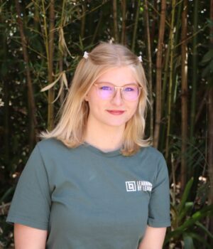 A person with blonde hair, glasses, and a green "Learning by Leaving" t-shirt stands outdoors in front of tall plants and bamboo.