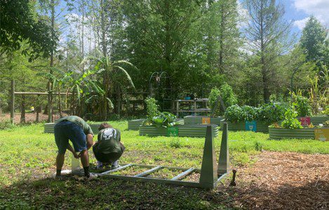 Two people are assembling a metal frame on the ground in a garden with raised beds containing plants, surrounded by trees and greenery.