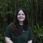 A woman with long brown hair stands in front of tall green plants, wearing a dark green T-shirt and smiling slightly at the camera.