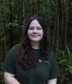 A woman with long brown hair stands in front of tall green plants, wearing a dark green T-shirt and smiling slightly at the camera.