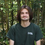 A person with shoulder-length brown hair sits outdoors in front of tall bamboo, wearing a dark green “Learning By Leading” T-shirt and smiling slightly.