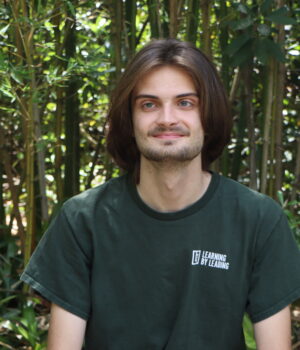 A person with shoulder-length brown hair sits outdoors in front of tall bamboo, wearing a dark green “Learning By Leading” T-shirt and smiling slightly.