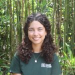 A young woman with curly hair sits on a bench in front of tall green plants, wearing a dark green “Learning & Leading” t-shirt and a silver necklace, smiling at the camera.