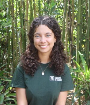 A young woman with curly hair sits on a bench in front of tall green plants, wearing a dark green “Learning & Leading” t-shirt and a silver necklace, smiling at the camera.