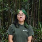 A person with green and black hair, glasses, and a green shirt stands in front of dense bamboo and leafy plants.