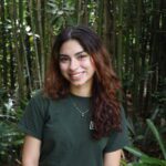 A person with long, wavy brown hair and a green shirt smiles while standing in front of tall bamboo and dense greenery.