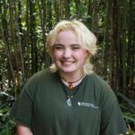 Person with short blond hair and a green shirt stands smiling in front of tall bamboo and leafy plants.