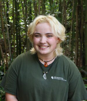 Person with short blond hair and a green shirt stands smiling in front of tall bamboo and leafy plants.