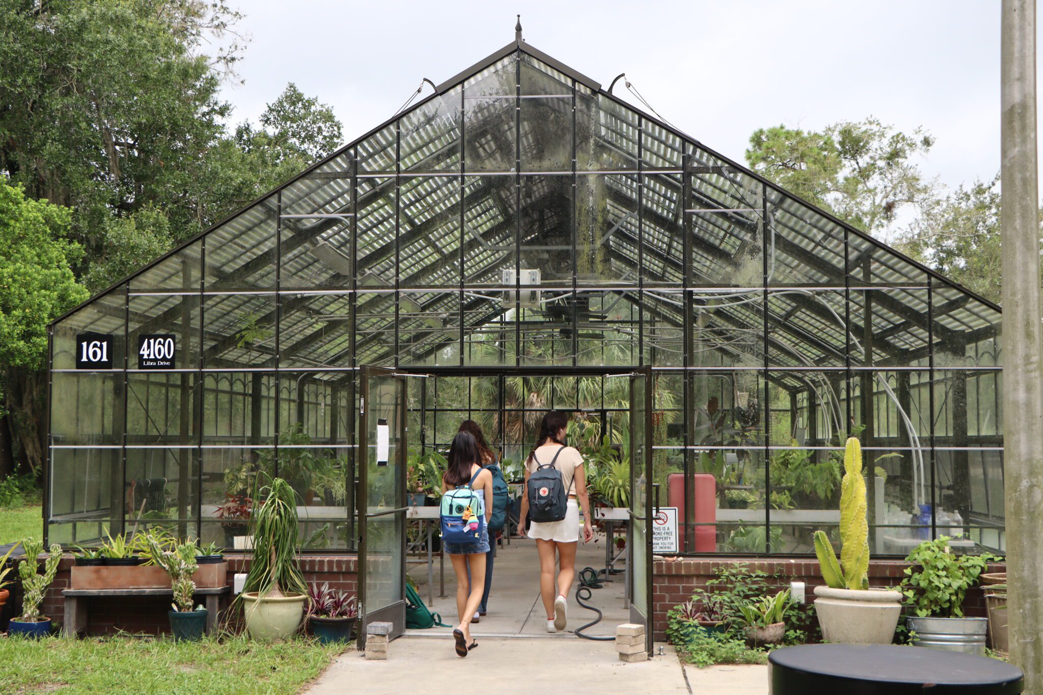 Two people with backpacks walking into a glass greenhouse with an address sign labeled 161 Libra Drive.